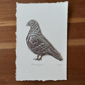 May include: A silver metal pigeon art piece on a white textured paper. The pigeon has a flower design on its body and the word "Pigeon" is written below it. The paper has a deckled edge.