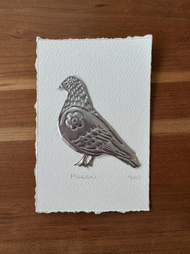 May include: A silver metal pigeon art piece on a white textured paper. The pigeon has a flower design on its body and the word "Pigeon" is written below it. The paper has a deckled edge.
