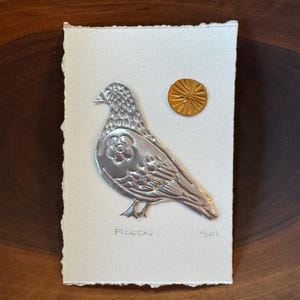 May include: A silver metal pigeon art piece on textured white paper, with a gold sun-like accent. The word "PIGEON" is written below the bird. The artwork is mounted on a wooden surface.