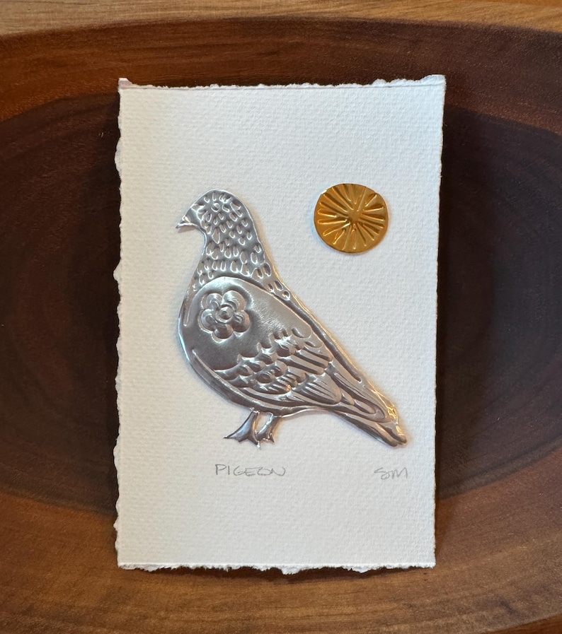 May include: A silver metal pigeon art piece on textured white paper, with a gold sun-like accent. The word "PIGEON" is written below the bird. The artwork is mounted on a wooden surface.
