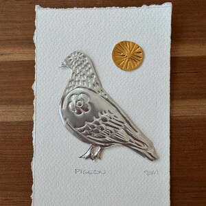 May include: A handmade artwork featuring a silver metal pigeon relief on textured white paper. A small gold-colored sun shape is above the bird. The word "PIGEON" is written below the bird.