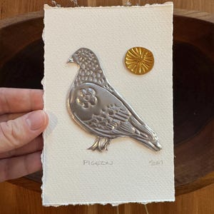 May include: A silver metal pigeon art piece on textured white paper, with a gold sunburst accent. The word "PIGEON" is written below the bird. The artwork is held by a hand.