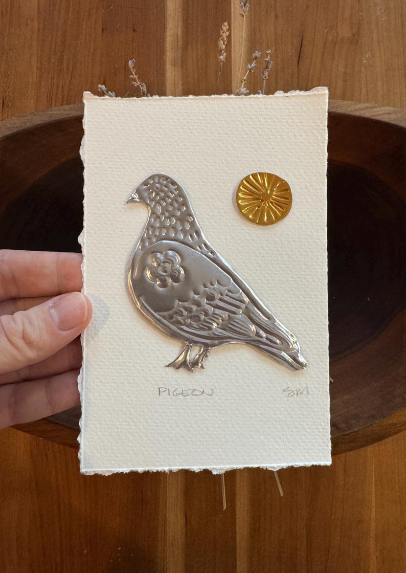 May include: A silver metal pigeon art piece on textured white paper, with a gold sunburst accent. The word "PIGEON" is written below the bird. The artwork is held by a hand.
