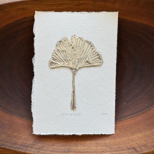 以下が含まれることがあります： 金属製のイチョウの葉の彫刻が、質感のあるオフホワイトの紙に固定されています。 葉には葉脈が詳細に描かれており、下に「GINKGO」の文字が書かれています。 紙は木製のボウルにセットされています。