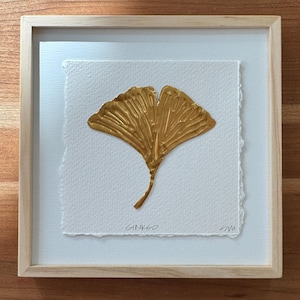 Puede incluir: Una obra de arte enmarcada con una hoja de ginkgo dorada sobre papel blanco texturizado. La hoja está pintada con pinceladas visibles. La palabra "GINKGO" está escrita en tinta negra debajo de la hoja. El marco es de madera clara.