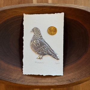 May include: A silver-colored pigeon relief art piece on a textured white paper, with a small gold sun-like embellishment. The word "Pigeon" is written below the bird. The artwork is set in a wooden bowl.