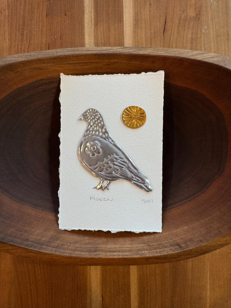 May include: A silver-colored pigeon relief art piece on a textured white paper, with a small gold sun-like embellishment. The word "Pigeon" is written below the bird. The artwork is set in a wooden bowl.