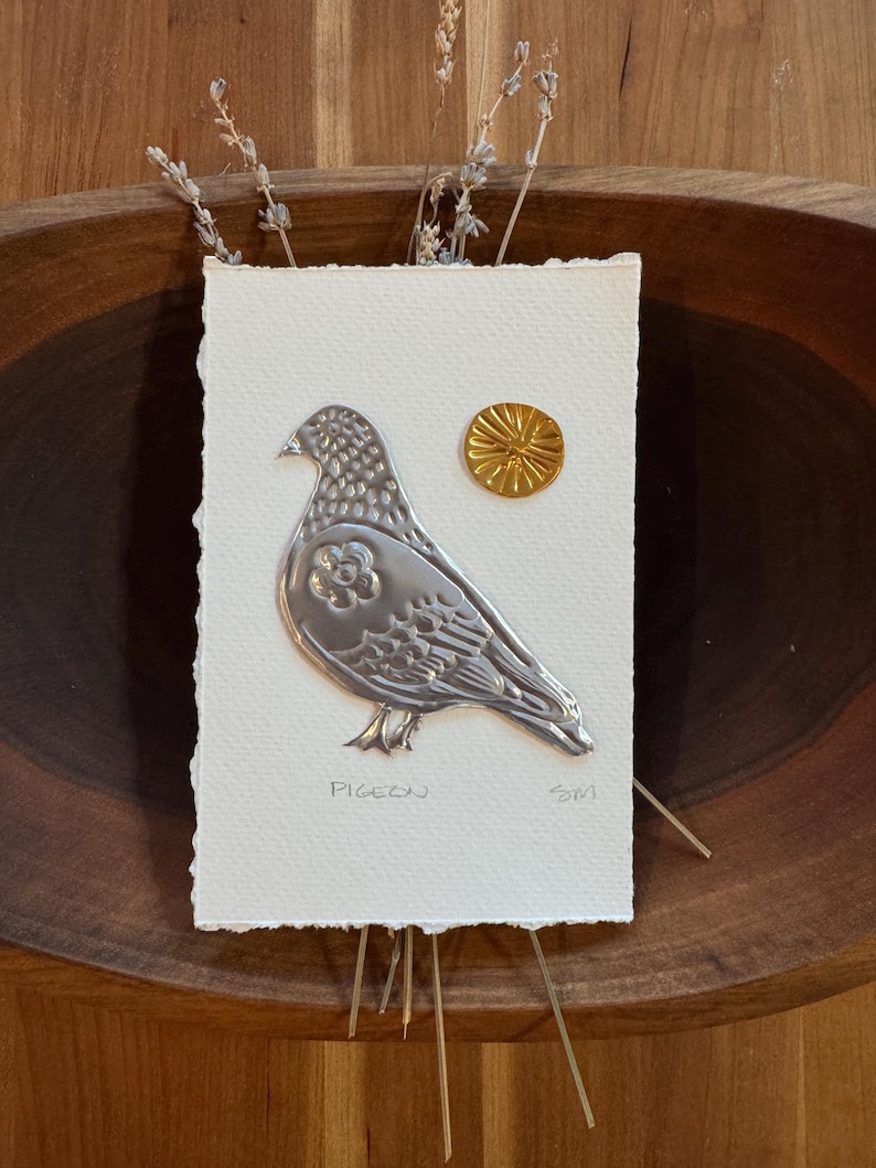 May include: A silver metal pigeon relief art piece on textured white paper, with a small gold sun-like embellishment. The word "PIGEON" is printed below the bird. Dried sprigs of lavender are placed above the art piece.
