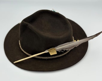 May include: A dark brown fedora hat with a wide brim. The hat is decorated with a gold-colored chain around the crown and a feather attached with a gold-colored metal piece. The feather is gray and brown.