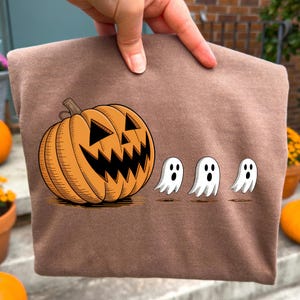 Pode incluir: Um moletom marrom com tema de Halloween com uma abóbora laranja esculpida e três ilustrações de fantasmas brancos. O desenho é impresso em tecido marrom.