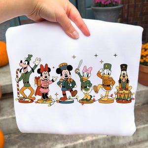 May include: White sweatshirt with a festive design featuring Disney characters in military-style outfits. Goofy holds a candy cane, Minnie Mouse has a gift, Mickey Mouse points, Daisy Duck holds a sword, Donald Duck salutes, and Pluto sits on a drum.