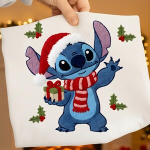 May include: A white pillow featuring a blue cartoon character wearing a Santa hat and a red and white striped scarf. The character is holding a wrapped gift. Embroidered holly sprigs are scattered around the image.