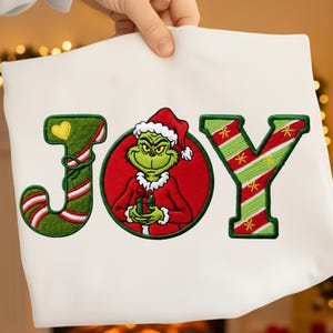 Könnte beinhalten: Weißer Stoff mit dem Wort "JOY" in Grün, Rot und Weiß gestickt. Der Buchstabe "O" zeigt den Grinch mit Weihnachtsmütze, der ein Geschenk hält. Die Buchstaben sind grün umrandet und haben ein festliches Design.
