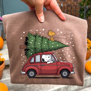 May include: Brown t-shirt featuring a cartoon Snoopy driving a red car with a Christmas tree on top. Woodstock is perched on the tree, with snowflakes falling around them. A festive holiday design.