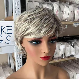 May include: A mannequin head with a short, layered wig in shades of gray and blonde. The mannequin has blue eyes, red lipstick, and dramatic eye makeup. A sign in the background reads "LAKE WIGS". Shelves in the background are filled with packaged items.