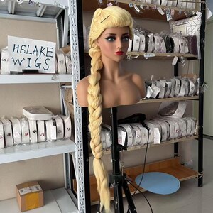 Pode incluir: Uma peruca loira com uma trança longa e grossa é exibida em uma cabeça de manequim. A peruca é estilizada com uma risca lateral e uma seção trançada. O manequim é montado em um suporte preto. A placa de fundo diz "HSLAKE WIGS".