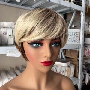 May include: A short, layered wig featuring a blend of blonde and light brown hair tones. The mannequin head displays a side-swept fringe, bright blue eyes, sharp black winged eyeliner, and bold red lipstick, presented in a storage environment.