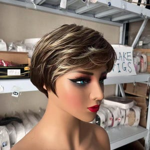 May include: A mannequin head with a short, layered wig in shades of brown and blonde. The mannequin has dramatic makeup with red lipstick and winged eyeliner. The background includes shelves with various items and a sign that reads "LAKE WIGS."
