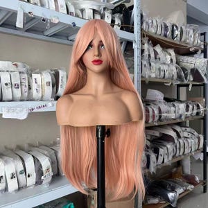 Puede incluir: Una peluca larga y lisa de color rosa pastel sobre una cabeza de maniquí beige. La peluca presenta una raya central con capas largas que enmarcan el rostro y caen más allá de los hombros. El maniquí tiene rasgos faciales pintados.