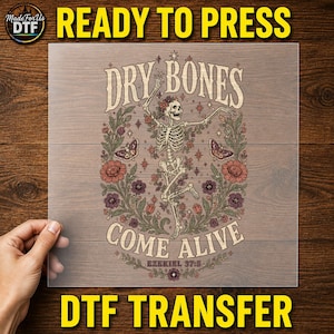 May include: A DTF transfer sheet featuring a skeleton dancing amidst colorful flowers, butterflies, and stars. The text reads DRY BONES COME ALIVE and EZEKIEL 37:5. The design uses a palette of muted red, purple, green, and cream tones on a clear film.