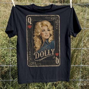 May include: A black short-sleeve t-shirt featuring a graphic print designed like a playing card. The print shows a portrait of a blonde woman with voluminous hair, the text DOLLY, the letter Q, and red heart symbols in a vintage style.