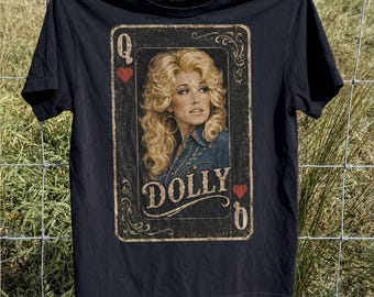 May include: A black short-sleeved t-shirt featuring a graphic print designed like a playing card. The print shows a portrait of a blonde woman with voluminous hair, the text DOLLY, the letter Q, and red heart symbols in a vintage style.