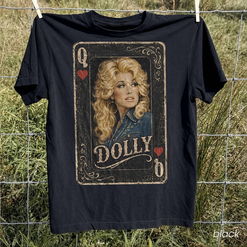 May include: A black short-sleeve t-shirt featuring a graphic print designed like a playing card. The print shows a portrait of a blonde woman with voluminous hair, the text DOLLY, the letter Q, and red heart symbols in a vintage style.