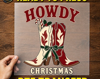 May include: A DTF transfer sheet featuring the text HOWDY CHRISTMAS in red and cream lettering. The design displays two cowboy boots, one cream and one red, tied together with a green ribbon. A candy cane and holly leaves are tucked into the cream boot.