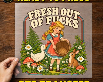 May include: A DTF transfer sheet featuring a cartoon illustration of a blonde girl in a blue and red dress holding a woven basket. The text reads FRESH OUT OF FUCKS. The scene includes a white duck, a brown rabbit, ferns, daisies, and red mushrooms.