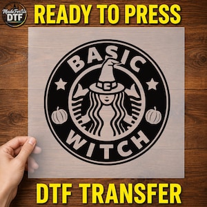 May include: A black DTF transfer sheet featuring a circular graphic design. The center shows a figure wearing a witch hat with long hair, surrounded by the text BASIC WITCH. Two small pumpkins and two stars are positioned within the circular border.