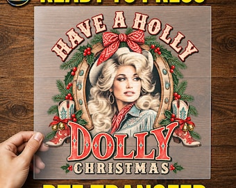 May include: A DTF transfer sheet featuring a graphic of a blonde woman in a cowboy hat and bandana, framed by a horseshoe, pine branches, red berries, and cowboy boots. The text reads HAVE A HOLLY DOLLY CHRISTMAS in red and white lettering.