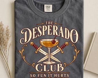 May include: A charcoal grey t-shirt featuring a graphic of a cocktail glass with a cherry, flanked by two crossed daggers. The text reads THE DESPERADO CLUB and SO FUN IT HURTS in a decorative font. The shirt has a crew neckline and a Comfort Colors label.