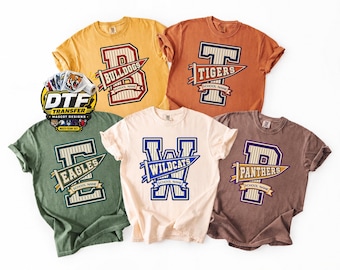 May include: Five short-sleeved t-shirts in yellow, orange, green, cream, and brown, each featuring a large varsity-style letter with a sports pennant graphic. The designs include the text Bulldogs, Tigers, Eagles, Wildcats, and Panthers.