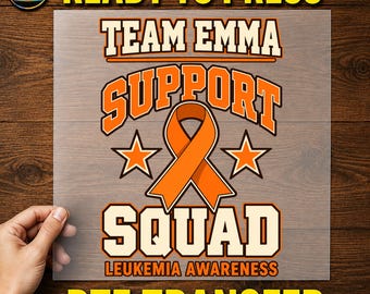 May include: A DTF transfer sheet featuring the text TEAM EMMA SUPPORT SQUAD LEUKEMIA AWARENESS in orange and white block lettering. The design includes a central orange awareness ribbon flanked by two orange stars on a clear film.
