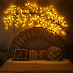 May include: A bedroom scene featuring a bed with a woven headboard and decorative pillows. Above the bed, a branch-shaped string light with warm yellow lights illuminates the wall. The overall aesthetic is cozy and inviting.