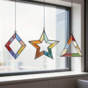 Può includere: Tre decorazioni per finestre in vetro colorato a forma di diamante, stella e triangolo. Ognuna ha una cornice in metallo nero ed è sospesa da un sottile filo nero. I pezzi di vetro sono di vari colori, tra cui rosso, blu, giallo e verde.