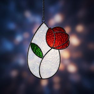 May include: Stained glass suncatcher in a teardrop shape, featuring a red rose and green leaves. The rose is made of red glass, and the leaves are green. The background is white, and the piece is outlined in black. A metal chain is attached for hanging.