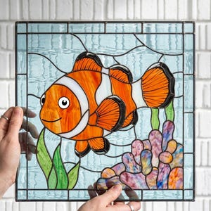 May include: A square stained glass panel featuring a vibrant orange and white clownfish swimming among green seaweed and multicolored coral. The background is composed of light blue glass pieces with dark lead lines forming a grid pattern.