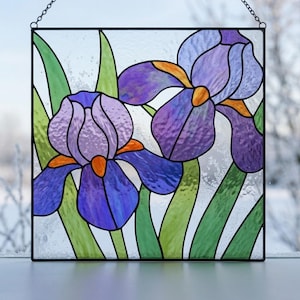 May include: A square stained glass panel featuring two purple iris flowers with orange centers and green leaves. The glass has a textured finish and is framed in black metal with a hanging chain attached to the top corners of the square frame.