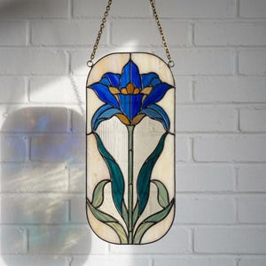 May include: An oval stained glass panel featuring a central blue flower with yellow accents, green leaves, and a cream-colored background. The piece is framed in dark metal and suspended by a gold-toned metal chain for hanging display purposes.