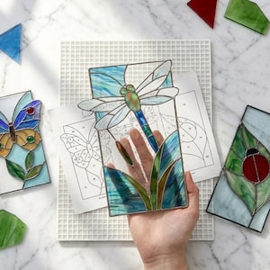 May include: Three rectangular stained glass panels featuring a dragonfly, a butterfly, and a ladybug. The dragonfly panel is held up, showing blue, green, and white glass pieces joined by metal solder lines, alongside paper patterns and loose glass.