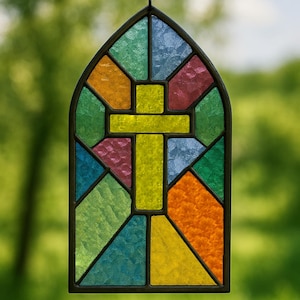 May include: A stained glass window with a cross design. The window features a variety of colors, including yellow, green, blue, orange, and red. The window is framed in a dark metal and is hanging from a chain.