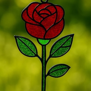 Puede incluir: Una rosa de vidriera con una flor roja y hojas verdes. La rosa está delineada en negro y cuelga de una cadena negra. El fondo es un verde suave y borroso.