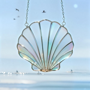 May include: A decorative stained glass seashell suncatcher. The shell is white with iridescent accents and a gold-colored metal trim. It hangs from a silver chain. The background is a blurred image of the ocean and sky.