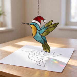 May include: A stained glass hummingbird suncatcher featuring blue, yellow, and green glass panels with a red and white Santa hat. The ornament hangs from a metal chain above a white paper template with numbered sections for assembly.