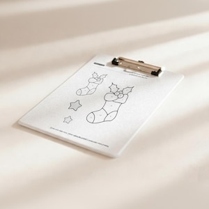 May include: A white clipboard with a silver clip holds a coloring page featuring Christmas stockings and stars. The stockings have holly accents and numbered sections for coloring. Text at the bottom reads, "If you want color, please adjust the printer's..."