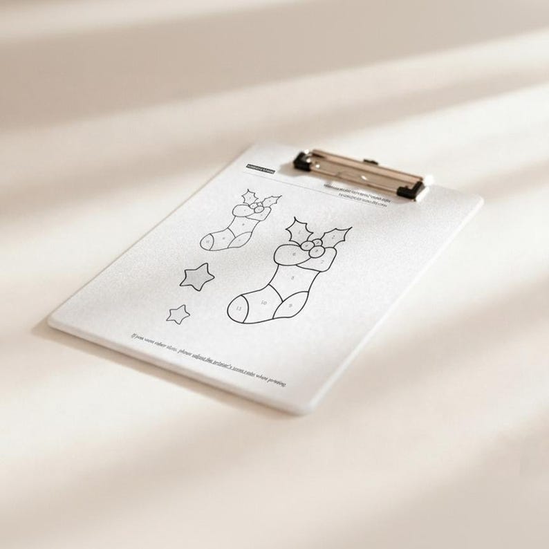 May include: A white clipboard with a silver clip holds a coloring page featuring Christmas stockings and stars. The stockings have holly accents and numbered sections for coloring. Text at the bottom reads, "If you want color, please adjust the printer's..."