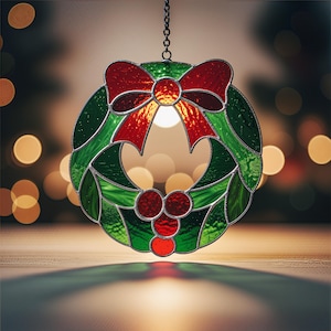 Op de afbeelding: Een gebrandschilderde kerstkrans met een rode strik en bessen. De krans is groen met rode accenten en hangt aan een ketting. De achtergrond is wazig met warme, onscherpe lichten.