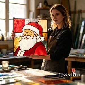 May include: A square stained glass panel featuring a depiction of Santa Claus with a white beard, mustache, and a red hat. The piece is constructed with various textures of red, white, and amber glass, framed by a thin metal border with two loops.