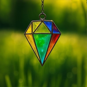 Puede incluir: Un colgante de vidrio coloreado en forma de diamante, con colores del arcoíris: amarillo, verde, azul y rojo. Cuelga de una cadena de metal oscuro sobre un fondo verde borroso.